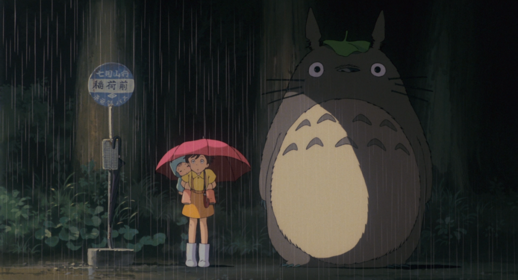 un frame del film animato Il mio vicino Totoro, con i protagonisti sotto la pioggia mentre attendono il bus 